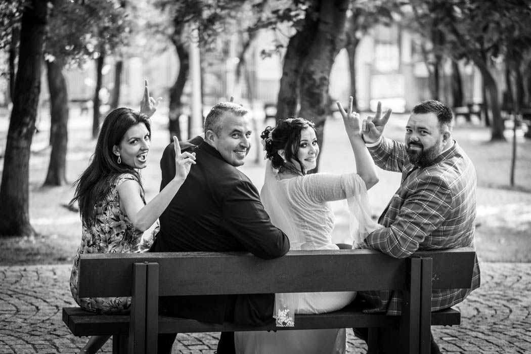 mirii si nasii pe banca in parc cu fundal blurat, intorsi spre fotograful de nunta, fac semnul victoriei